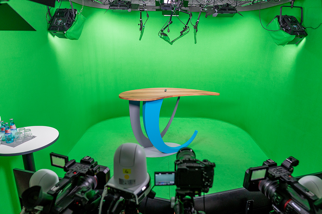 Der Regieraum zeigt mehrere Monitore mit Live-Bild aus einem Greenscreen-Studio. Mischpulte, ein Bildmischer, Audiotechnik, ein PTZ-Controller und weitere Geräte stehen auf dem Tisch. Auf den Monitoren sind zwei Personen im Studio zu sehen.